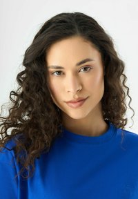 T-shirt en coton bleu avec un col rond ; présente un logo brodé sur la poitrine gauche ; le modèle a des cheveux bouclés et un maquillage neutre.