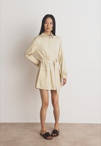 Béžové košeľové šaty s opaskom, dlhými rukávmi, golierom a dvojicou predných vreciek, skombinované s tmavohnedými šľapkami. Hladká textúra látky.