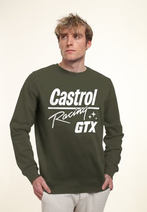 Ung mand iført en olivengrøn sweatshirt med hvid tekst "Castrol Racing GTX", der kigger let til siden med hænderne i lommerne.