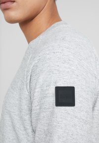 Šedá mikina s texturovanou tkaninou, kulatým výstřihem a černým čtvercovým štítkem s nápisem "BOSS" na levém rameni.