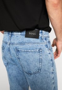Homme portant un jean en denim bleu clair avec une étiquette noire "BOSS" sur la ceinture arrière et une chemise noire rentrée à l'intérieur.