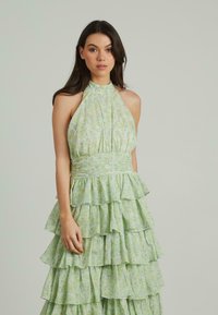 Zielona kwiatowa sukienka z dekoltem halter, warstwami falban i teksturowanym materiałem. Posiada delikatny wzór liści w jasnych odcieniach zieleni i żółci.