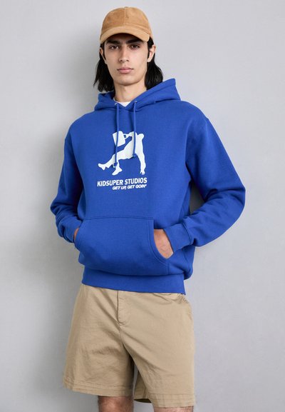Felpa blu in cotone con tasca frontale e stampa grafica bianca. Abbinata a pantaloni corti khaki e un cappellino beige.