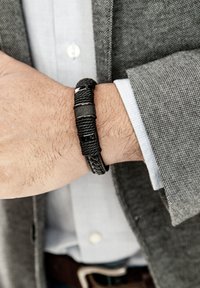 Schwarzes geflochtenes Armband mit verschiedenen Texturen und metallischen Akzenten, am Handgelenk befestigt und teilweise von einem grauen Blazer verdeckt.