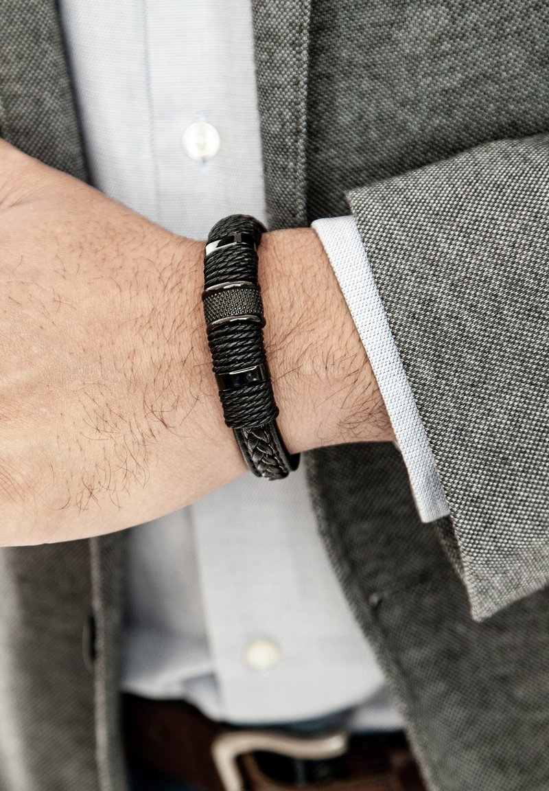 Schwarzes geflochtenes Armband mit verschiedenen Texturen und metallischen Akzenten, am Handgelenk befestigt und teilweise von einem grauen Blazer verdeckt.