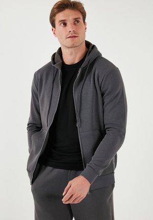 Un sweat à capuche gris zippé avec une texture douce, doté d'une capuche, de poches avant et d'un doublure intérieure noire contrastante, porté sur un t-shirt noir.