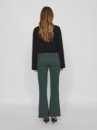 Un chandail noir à manches longues porté avec un pantalon évasé vert foncé. La tenue présente une texture lisse, avec des lignes simples et épurées ainsi qu'une silhouette ajustée.