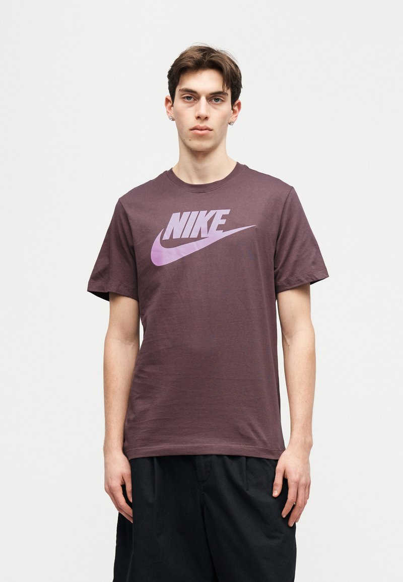 Jonge man met een paarse Nike T-shirt en zwarte korte broek, staand tegen een effen lichte achtergrond, recht vooruit kijkend.