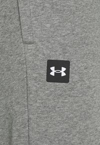Szary materiał bluzy z gładką teksturą. Zawiera czarne kwadratowe logo z białym stylizowanym emblematem "Under Armour".