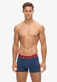 Marineblauwe boxershorts met een rode elastische tailleband waarop het logo "SUPERDRY" staat. Strakke pasvorm, zachte textuur en minimale naden.