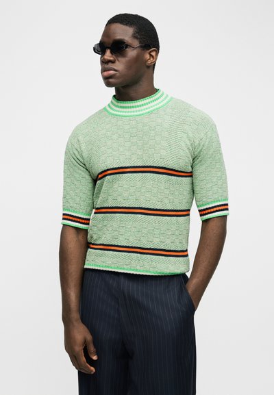 Muž nosiaci zelený textúrovaný krátky sveter s oranžovými a čiernymi pruhmi, tmavé nohavice s jemnými pásikmi a čierne slnečné okuliare, pozerajúci sa doprava.