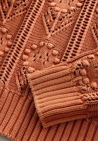 Tissu tricoté couleur rouille avec motifs bouclés et dentelle texturés, avec détail de poignet côtelé.