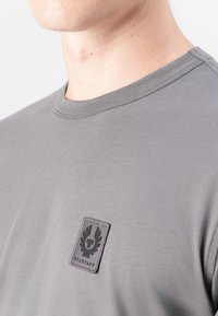 Tricou din bumbac gri, cu guler rotund și mâneci scurte. Are o etichetă logo de culoare gri închis pe partea stângă a pieptului. Textură netedă.
