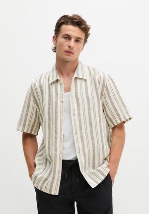 Jonge man met kort bruin haar, die een beige-wit verticaal gestreept kort mouwen overhemd draagt over een wit tanktop en zwarte broek.