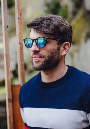 Černé sluneční brýle s modrými reflexními čočkami, kulatého tvaru, na muži s krátkým plnovousem, oblečeném v námořnickém a pruhovaném svetru.