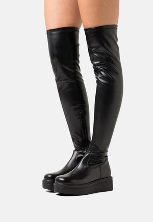 Botas negras hasta la rodilla hechas de cuero brillante, con una suela gruesa tipo plataforma y una cremallera lateral para facilitar su uso. Textura lisa por completo.