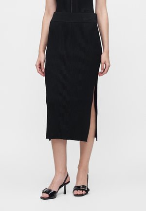 Kvinde iført en sort ribbet midi-nederdel med sideslids og sorte åbne tå-slingback-sandaler med spætte-detalje.