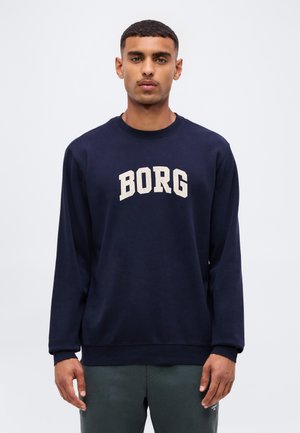 Jeune homme aux cheveux courts portant un sweat-shirt bleu marine avec le mot "BORG" en grandes lettres blanches, debout devant un fond clair uni.