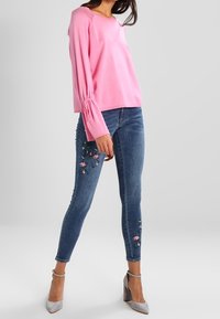Maglia rosa a maniche lunghe con polsini arricciati, abbinata a jeans skinny blu con ricami floreali rosa sulle gambe, e scarpe grigie con tacco alto.