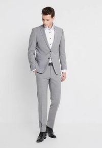 Szary garnitur o dopasowanym kroju z subtelnym wzorem w kratę, w połączeniu z białą koszulą z czarnymi guzikami i czarnymi butami.