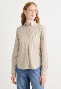 Camicia a righe nei toni beige e crema, con maniche lunghe, colletto e orlo curvo. Il tessuto appare liscio e leggero.