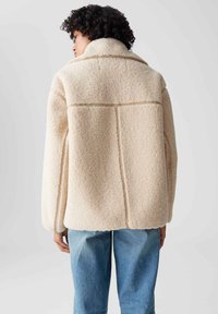 Cremfarbene, fuzzy Fleecejacke mit weitem Kragen, abgesenkten Schultern und gestickten Details. Kombinieren Sie sie mit hellblauem Denim.