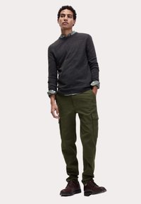 Donkergrijze trui, groene cargobroek met zijzakken, bruine veterschoenen en een lichtgroen overhemd met kraag eronder.