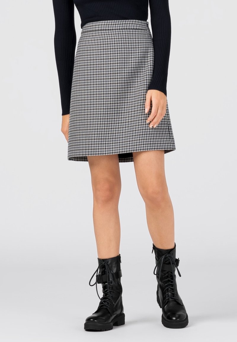 Mujer con camiseta negra de manga larga, falda de pata de gallo en blanco y negro, y botas negras de combate con cordones, de pie sobre un fondo liso.