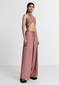 Donna in piedi che indossa pantaloni larghi a coste color malva con elastico e un top corto a halter abbinato, abbinati a sandali con plateau neri.