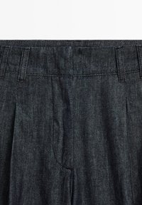 Pantalons en denim foncé avec un devant plissé, des passants de ceinture et des poches latérales. Le tissu a une texture fine et un subtil éclat.