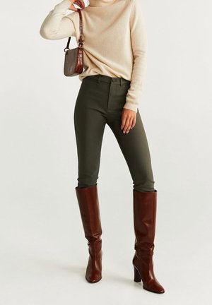 Frau trägt beigefarbenen Rollkragenpullover, dunkelgrüne eng anliegende Hose, braune kniehohe Lederstiefel und trägt eine kleine braune Umhängetasche.