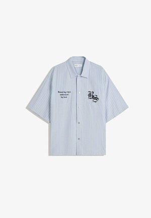 Camicia azzurra a maniche corte con bottoni e righe bianche verticali. Presenta testo e logo ricamati in nero sul lato sinistro.