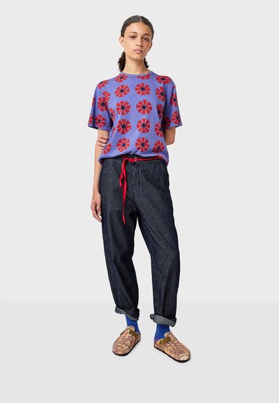 Fialové tričko s červeným květinovým vzorem, volné pruhované kalhoty s červeným šňůrkovým opaskem a barevné vzorované dřeváky.