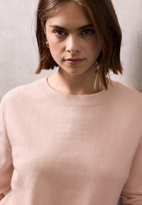 Jeune femme aux cheveux bruns courts portant un pull léger rose, se tenant devant un mur clair, regardant droit devant elle.