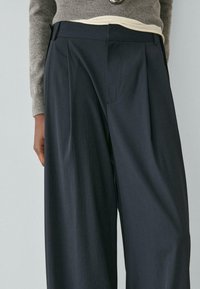 Dunkelblaue, weit geschnittene Hose mit plissiertem Vorderteil, glatter Textur und verdecktem Bund, kombiniert mit einem hellen Oberteil darunter.