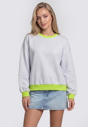 Jeune femme aux cheveux blonds clairs porte un sweat-shirt gris clair avec des poignets et un col vert néon, ainsi qu'une mini-jupe en denim bleu clair.