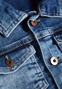 La giacca di jeans presenta un colore blu sbiadito, colletto classico, due tasche sul petto con bottoni metallici marroni e dettagli di cucitura visibili.