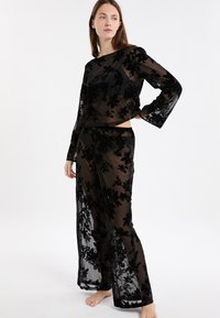 Vrouw in sheer zwart bloemenpatroon met lange mouwen en bijpassende wijde broek, staand op blote voeten tegen een witte achtergrond.