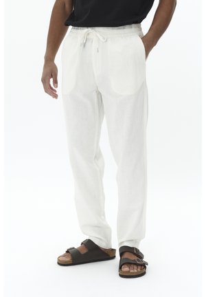 Hombre que lleva pantalones blancos sueltos con cordón, camiseta negra y sandalias negras de doble tira, de pie con las manos relajadas.