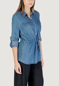 Camicia di jeans blu medio, con vita sagomata e un nastro, chiusura con bottoni davanti e due tasche sul petto. Maniche lunghe, arrotolate ai polsini.