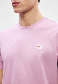 Lys rosa bomull-t-skjorte med rund hals, korte ermer og en liten, firkantet Calvin Klein-logo på brystet. Glatt tekstur.
