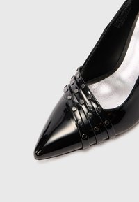Pantofii cu toc, din piele patent neagră, cu vârf ascuțit și patru bretele cu ținte pe partea frontală, având o căptușeală interioară din metalic argintiu.