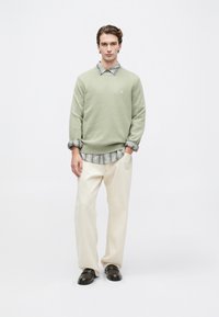 Jovem vestido com um suéter verde claro sobre uma camisa riscada cinza, calças crema e sapatos marrons escuros, de pé com uma mão no bolso.