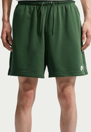 Mann trägt dunkelgrüne Nike-Sportshorts mit Kordelzug in der Taille und steht mit entspannten Händen an den Seiten vor einem einfarbigen Hintergrund.