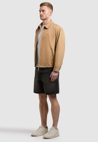 Jeune homme portant une veste à fermeture éclair beige, un t-shirt gris clair, un short en denim noir et des baskets blanches, debout devant un fond clair uni.