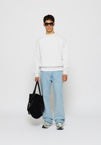 Young man wearing black sunglasses, white sweatshirt, light blue jeans, and sneakers, holding a black tote bag against a plain white background.