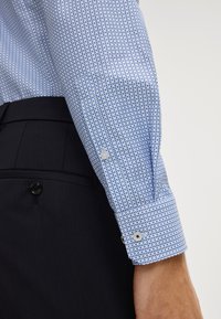 Camicia azzurra chiara con piccoli pois blu, dotata di bottoni bianchi e polsini alla francese, abbinata a pantaloni sartoriali blu navy scuro.