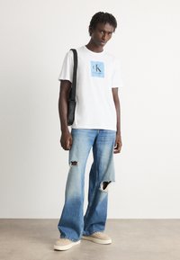 Balta marškinėliai su mėlynu Calvin Klein logotipu, derinami su šviesiai mėlynomis plačiomis kelnėmis su skylėmis keliuose ir kreminiais batais.