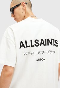 Fehér pamut póló, amelyen feltűnő fekete felirat található: "ALLSAINTS," japán karakterek, valamint "LONDON" a hátoldalon. Kerek nyakú, rövid ujjú.