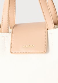 Sac à main texturé blanc avec des accents rose clair. Présente un logo "ALDO" doré sur un rabat rose et des poignées assorties. Fermeture zippée sur le dessus.